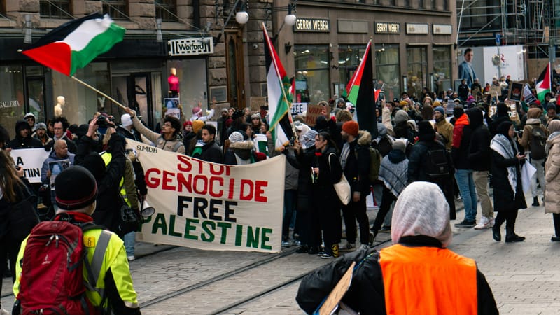 A group of people protesting, taking over the streets while holding Palestinian flags and a sig